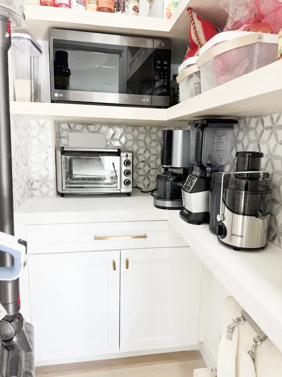 A corner pantry with white cabinets, a microwave on a shelf, and various countertop appliances against a geometric tile.