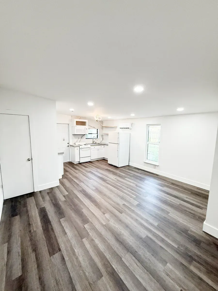 A bright, empty room with light gray wood-look flooring, white walls, recessed lighting, and a small kitchen area.