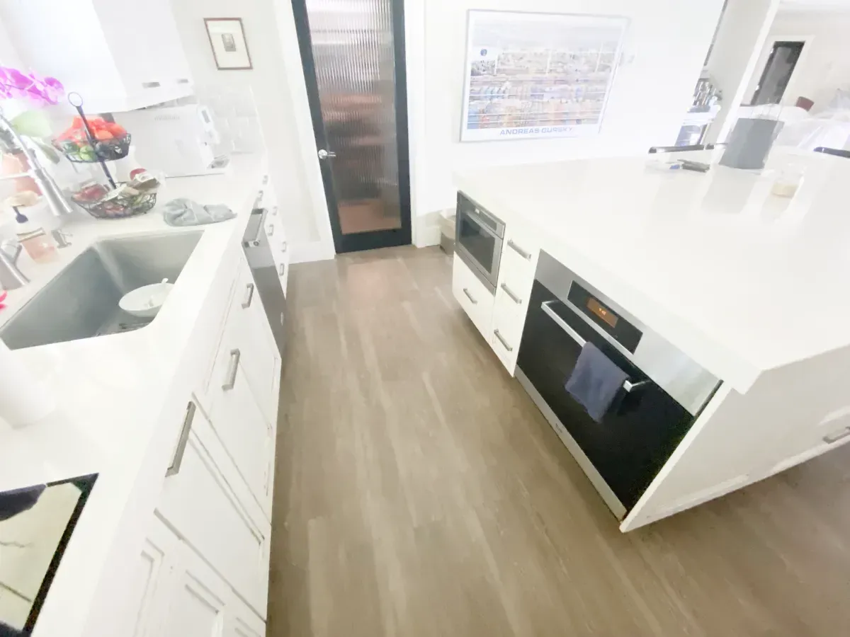 A bright modern kitchen with white countertops and cabinets, a stainless steel oven, and light wood-tone flooring.