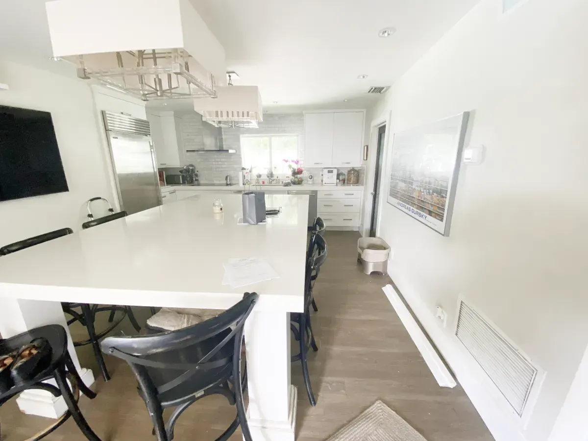 A bright, modern kitchen featuring a large white island with black chairs, leading into a galley-style cooking area.
