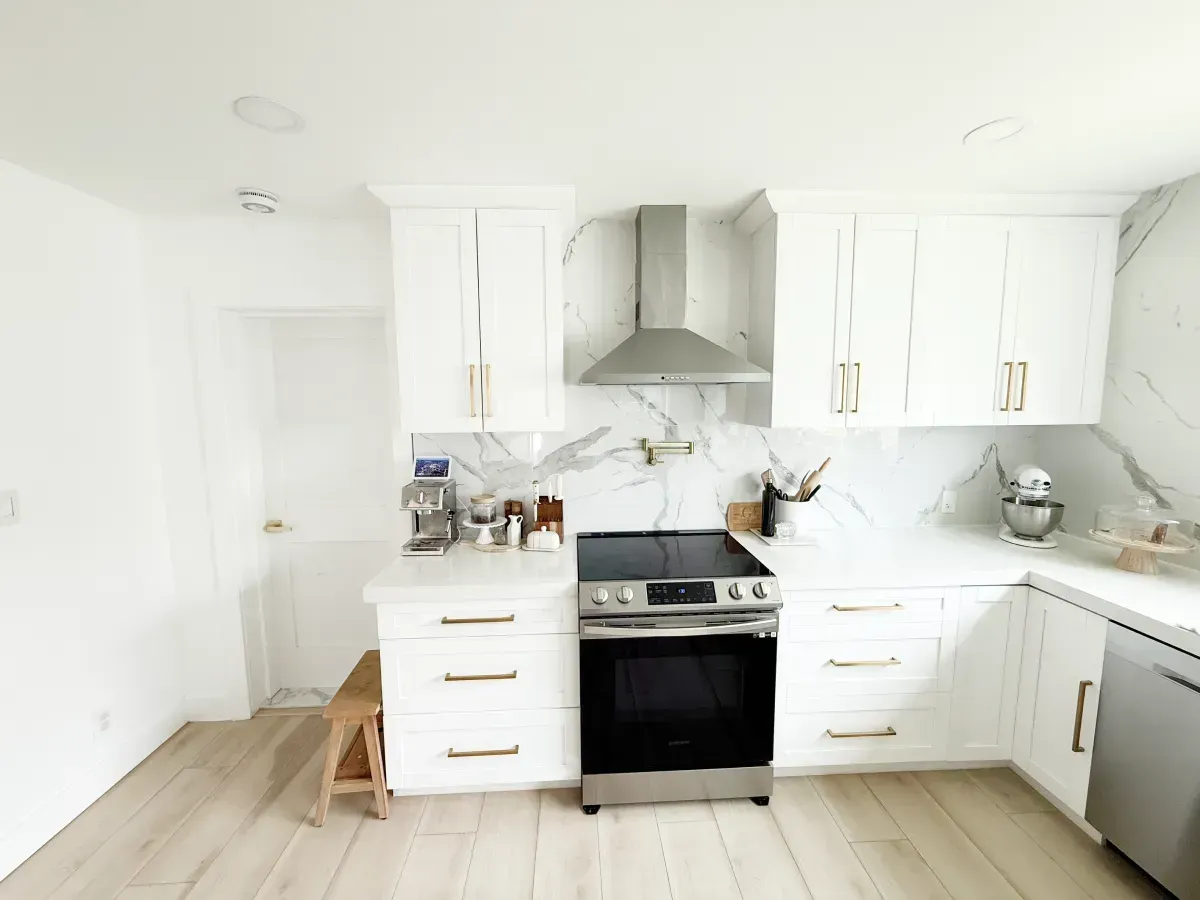 A modern kitchen with white cabinets, marble backsplash, light wood floors, and stainless steel appliances.