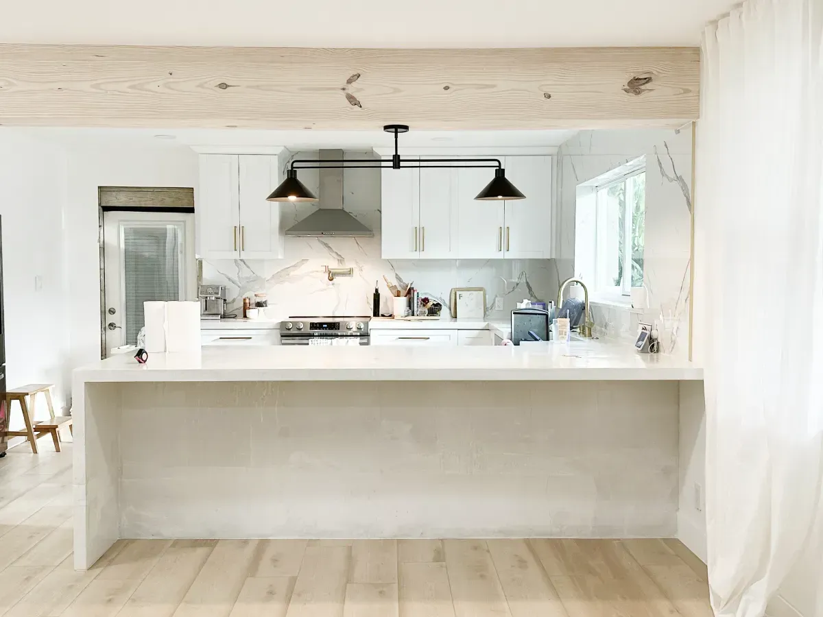 A bright, modern kitchen featuring white cabinets, a stone island, light wood flooring, and a decorative wood ceiling beam.