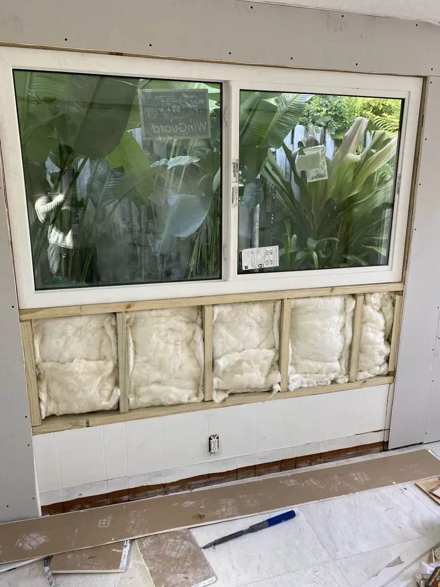 A wall section below a window with exposed wooden studs, fiberglass insulation, and a single electrical outlet.