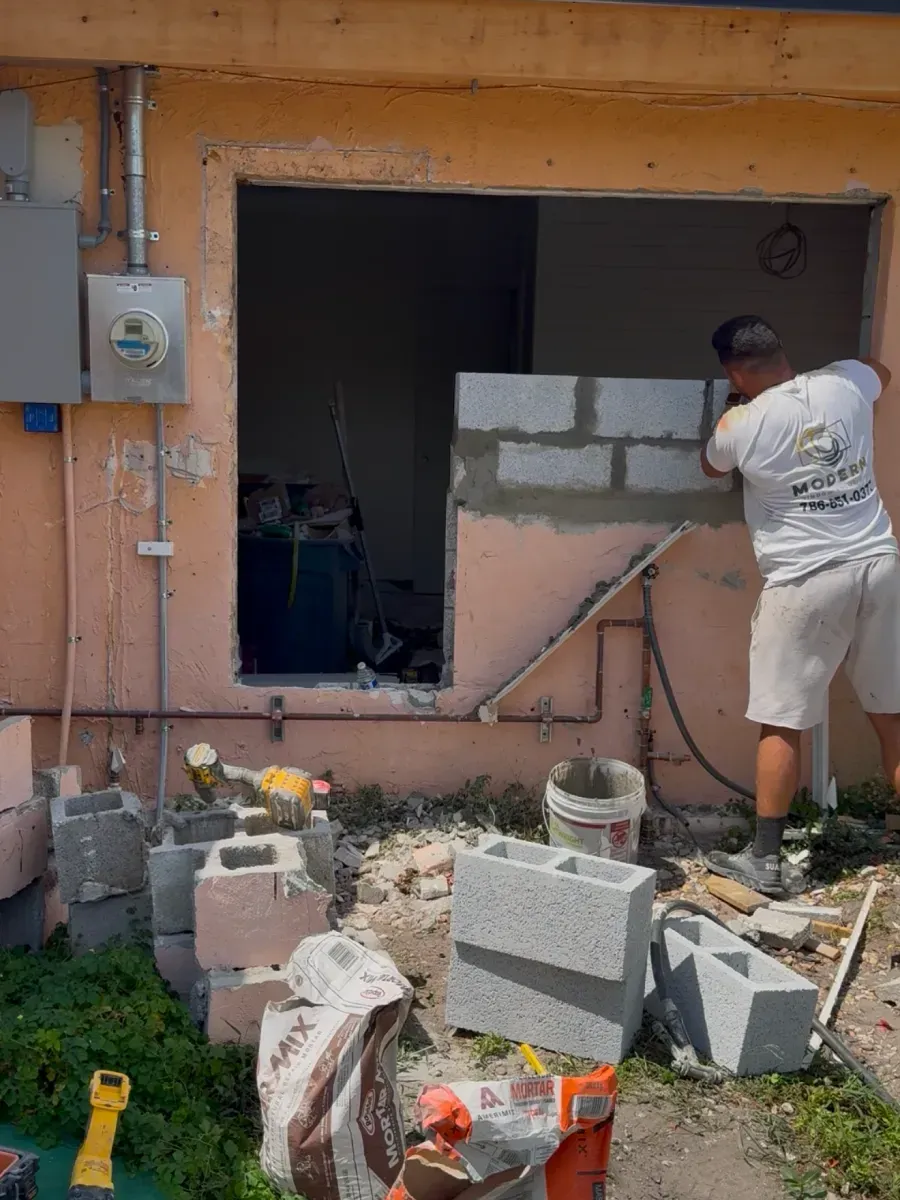 A construction worker uses concrete blocks to partially fill a large opening in an exterior peach-colored wall.