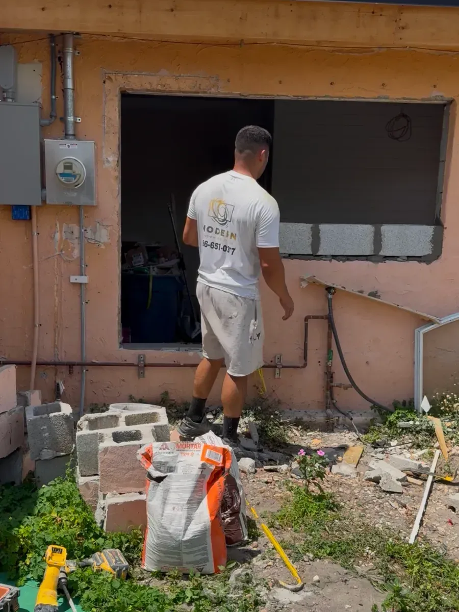 A person in a white t-shirt and light shorts works on a construction project involving a wall opening on a peach-colored house.
