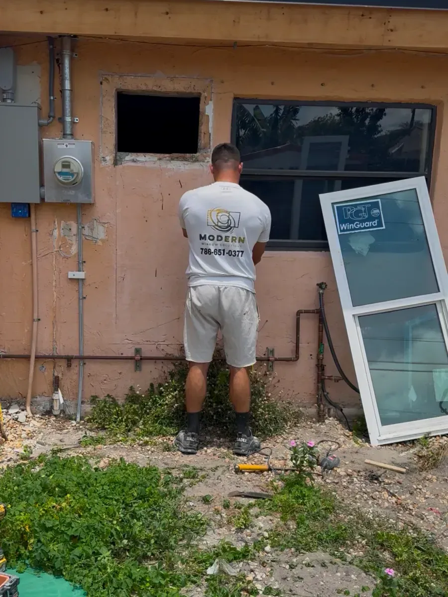 A person stands against a beige exterior wall, working on an open window frame, with a spare window unit nearby.