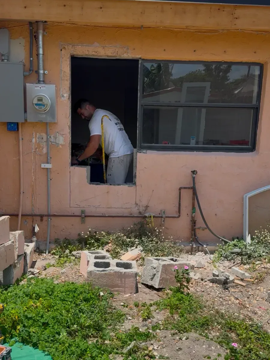 A person in a white t-shirt leans into a rectangular hole cut into the exterior wall of a peach-colored building.