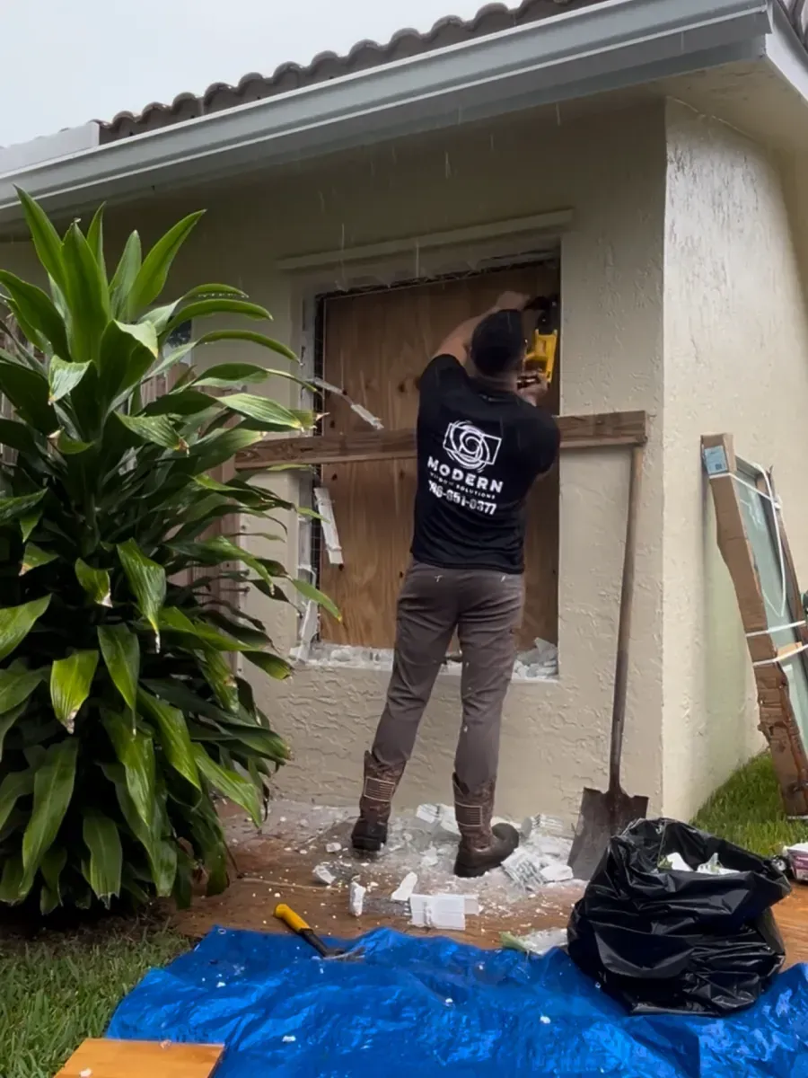 A person wearing a dark shirt works on removing a window frame from a stucco wall, with tools and debris on the ground.