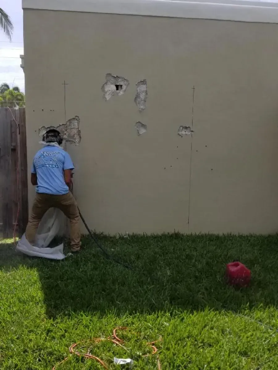 A person in a blue shirt uses a handheld tool to repair small holes in an exterior tan wall.