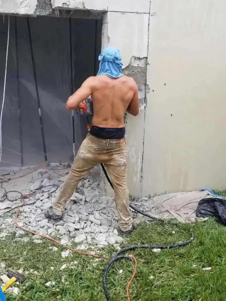 A shirtless worker wearing a blue head covering uses a power saw to cut an opening in a concrete wall.