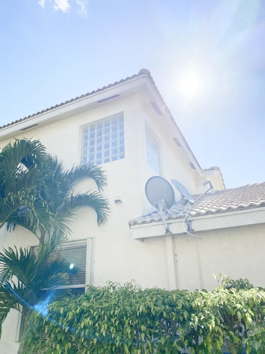 A low-angle view of a beige house exterior with a glass-block window, satellite dishes, a palm tree, and a green hedge.