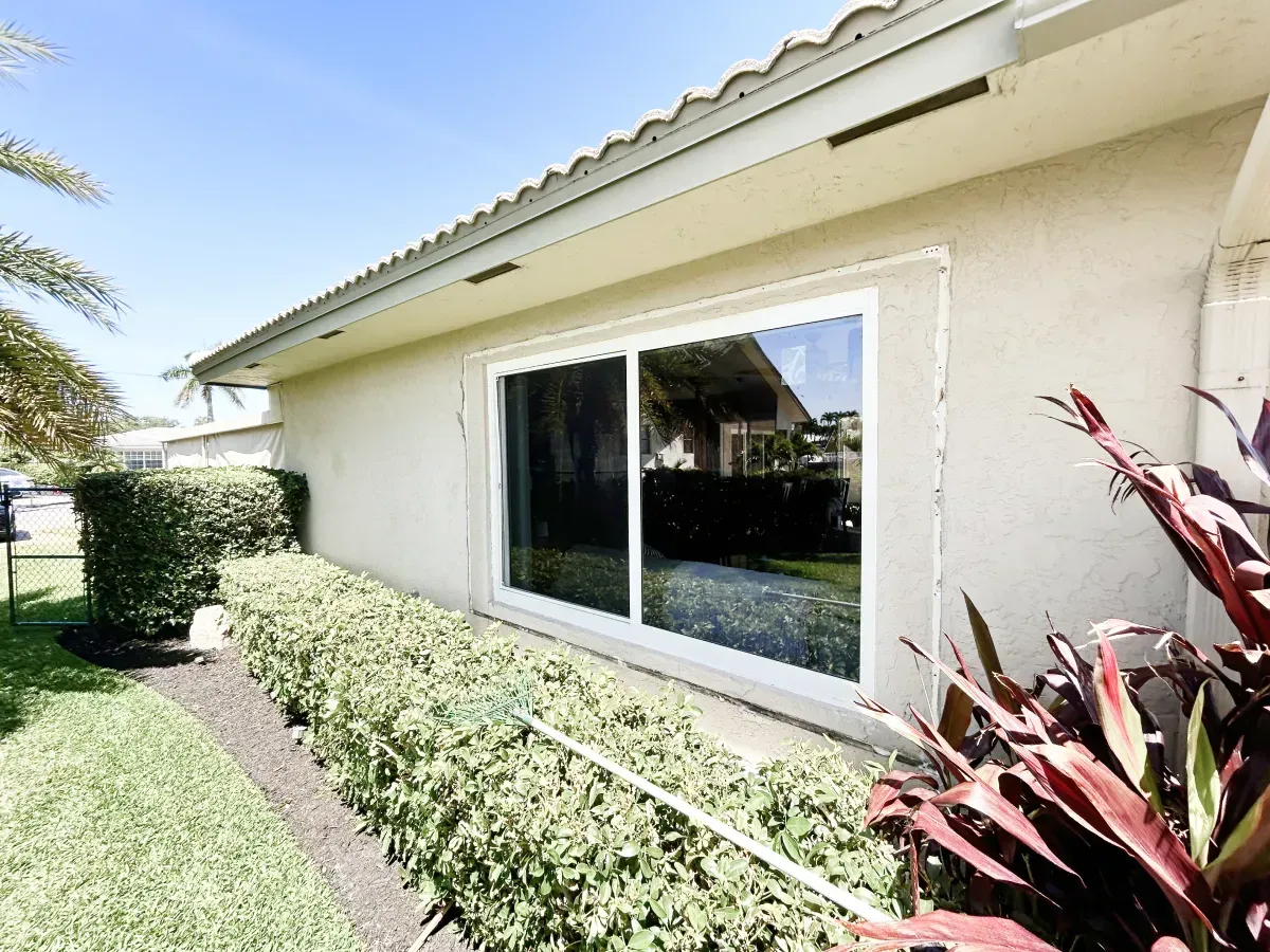 A side view of a cream-colored stucco home featuring a large white-framed sliding window above a manicured hedge.