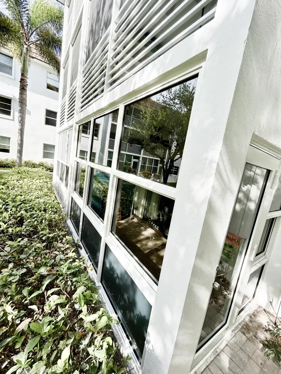 A white modern building exterior featuring rows of windows and louvered vents, situated next to green hedges.