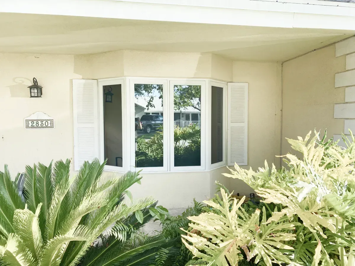 A bay window with white shutters on a beige house, partially obscured by lush green plants in the foreground.