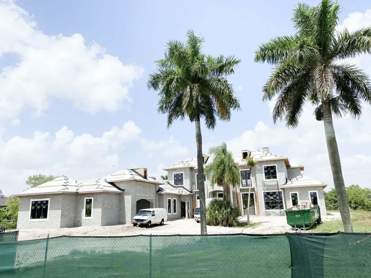 A large, light-colored house under construction, surrounded by palm trees and a green construction fence on a sunny day.