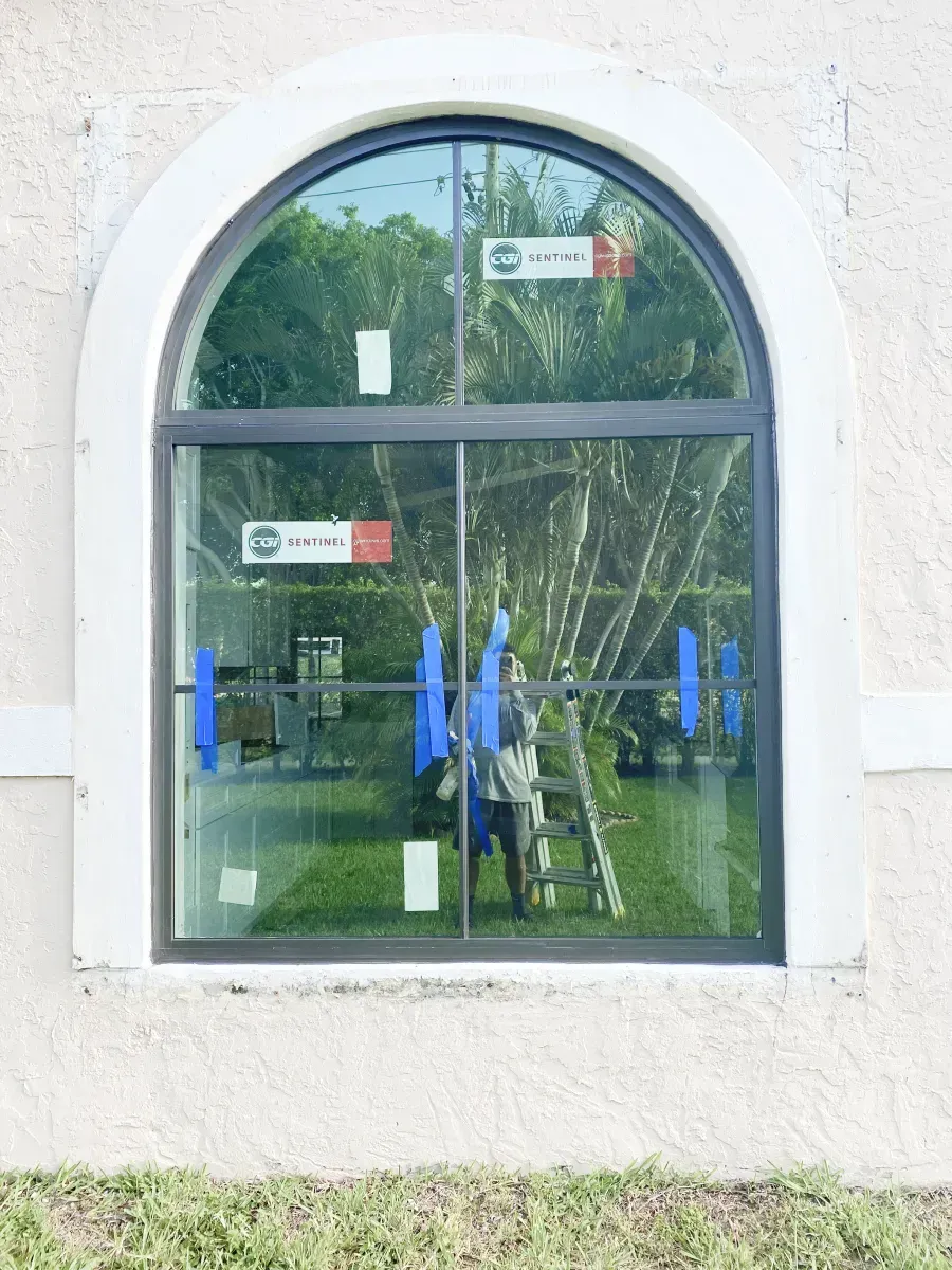 A dark-framed, arched window on a beige stucco wall, featuring blue painter's tape and manufacturer stickers on the glass.