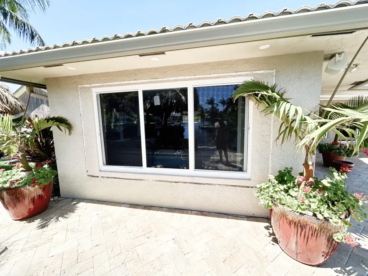 A beige stucco exterior wall with a three-pane white window, flanked by two large potted plants on a stone patio.
