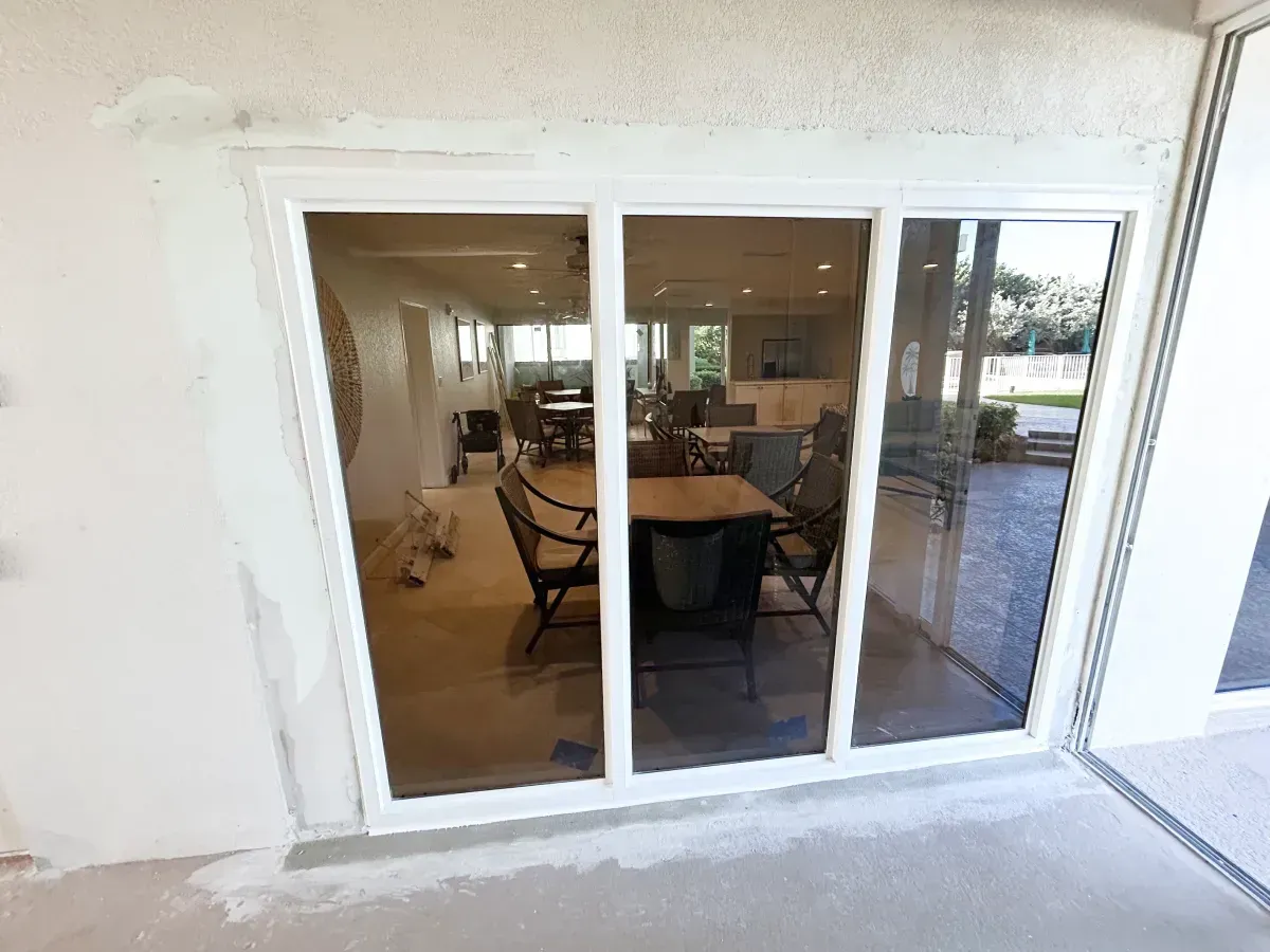 White sliding glass doors installed in a beige stucco wall, looking into a furnished patio dining area.
