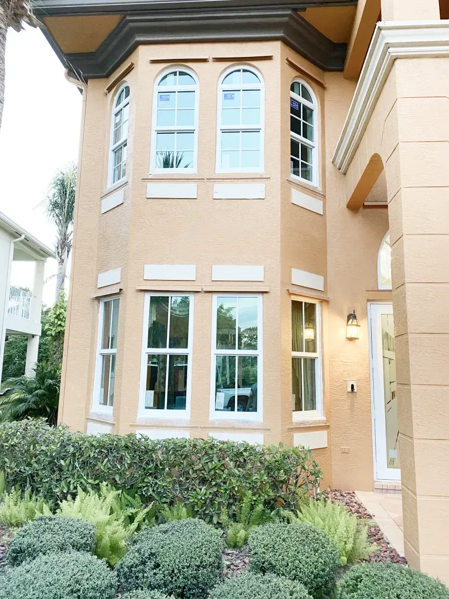 A tan, multi-story stucco house featuring arched upper windows and rectangular lower windows above a manicured hedge.