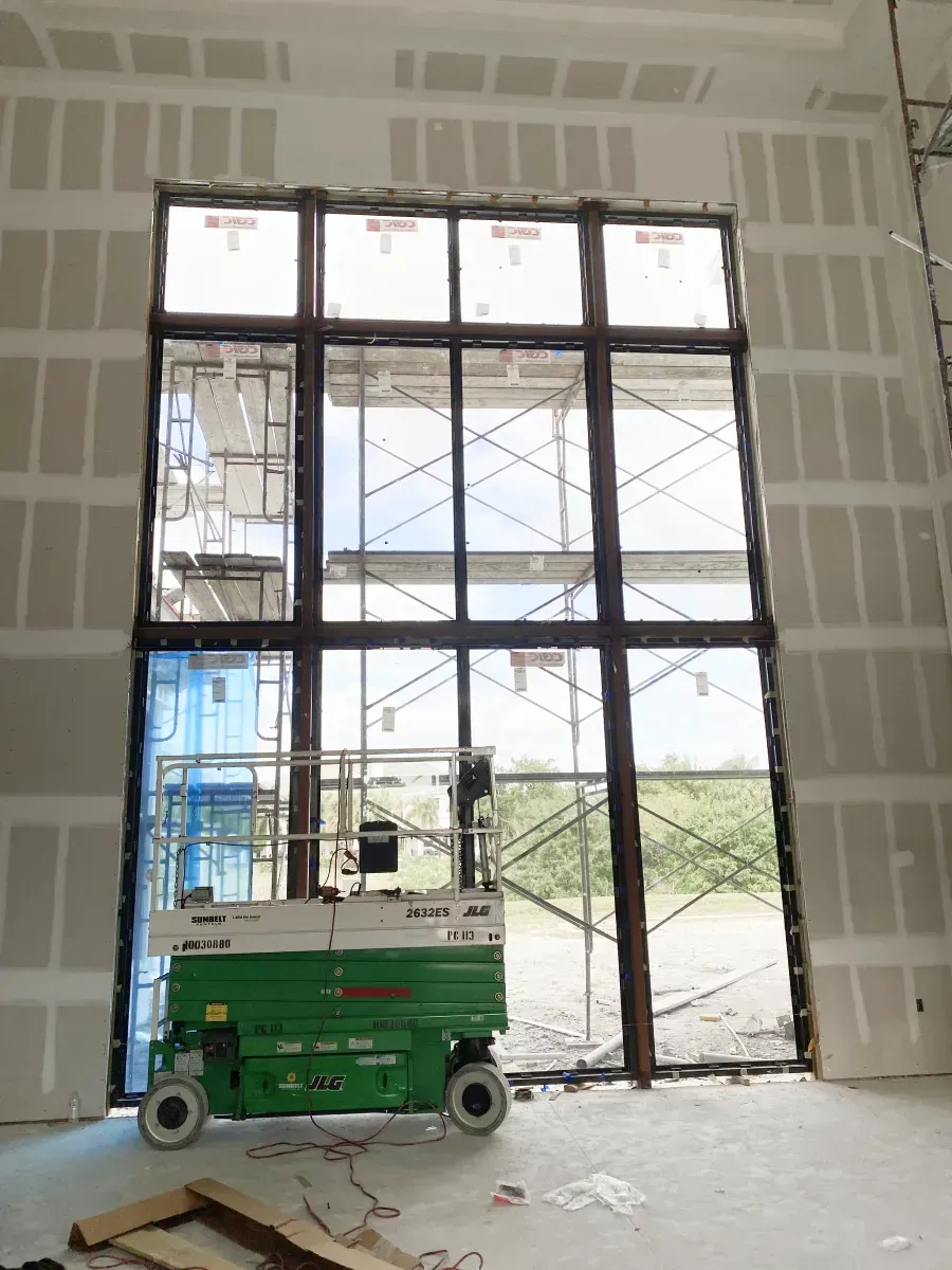 A green scissor lift sits inside a building under construction, parked before a large, multi-paned glass window.