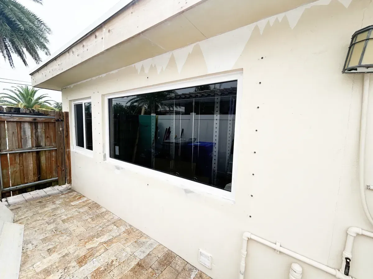 A light-colored exterior wall with two windows, a wooden fence on the left, and a paved patio area.