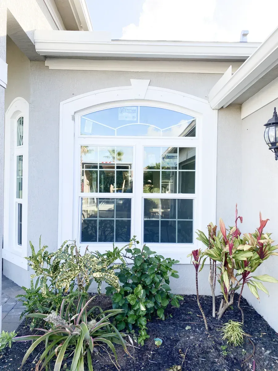 A white-framed arched window on a beige house exterior, featuring decorative grid patterns and a garden in front.