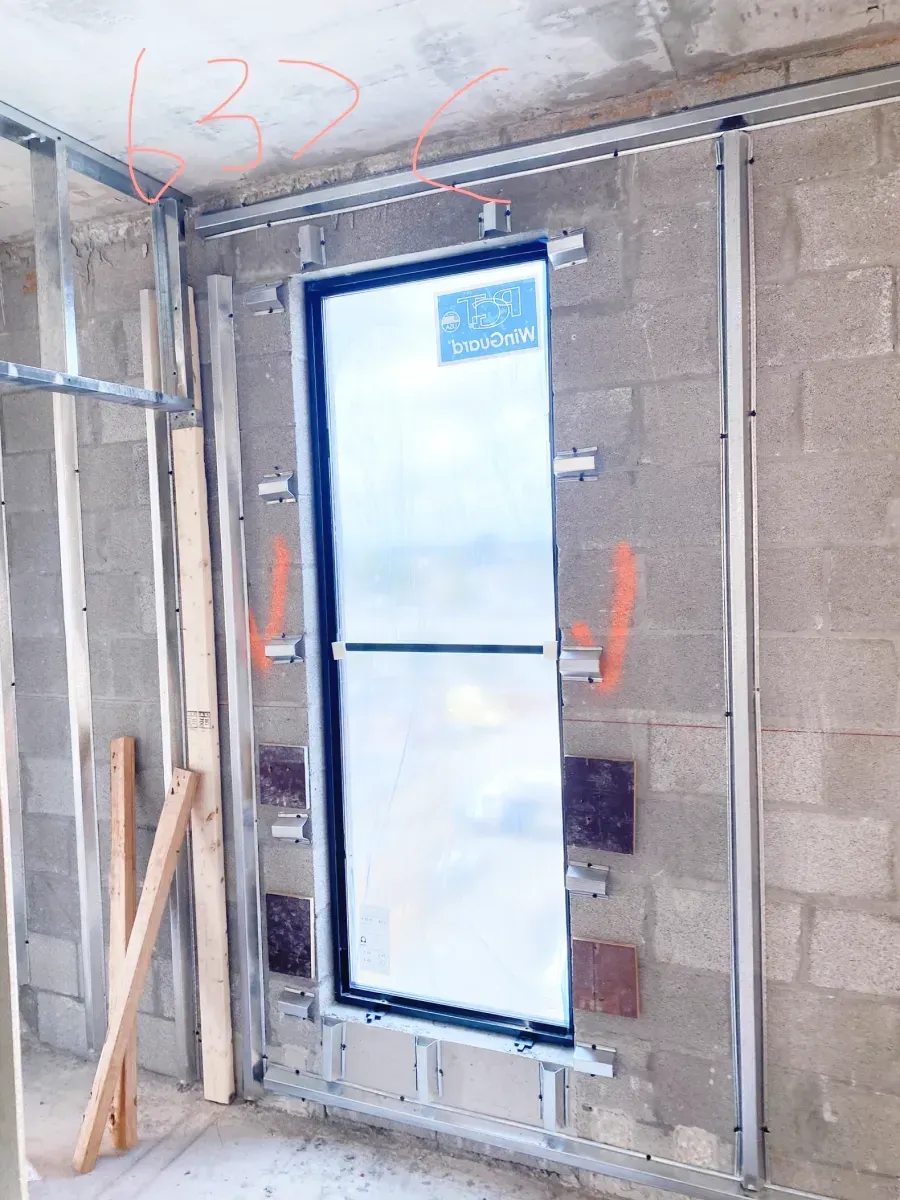 A vertical, framed window installed in a cinder block wall during construction, featuring metal tracks and orange marks.