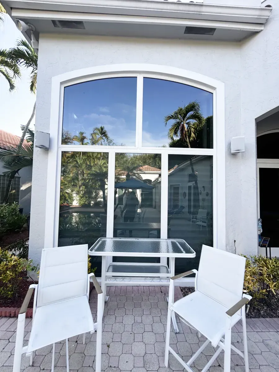 Two white chairs and a glass-topped table on a stone patio in front of a tall, arched window of a light-colored house.