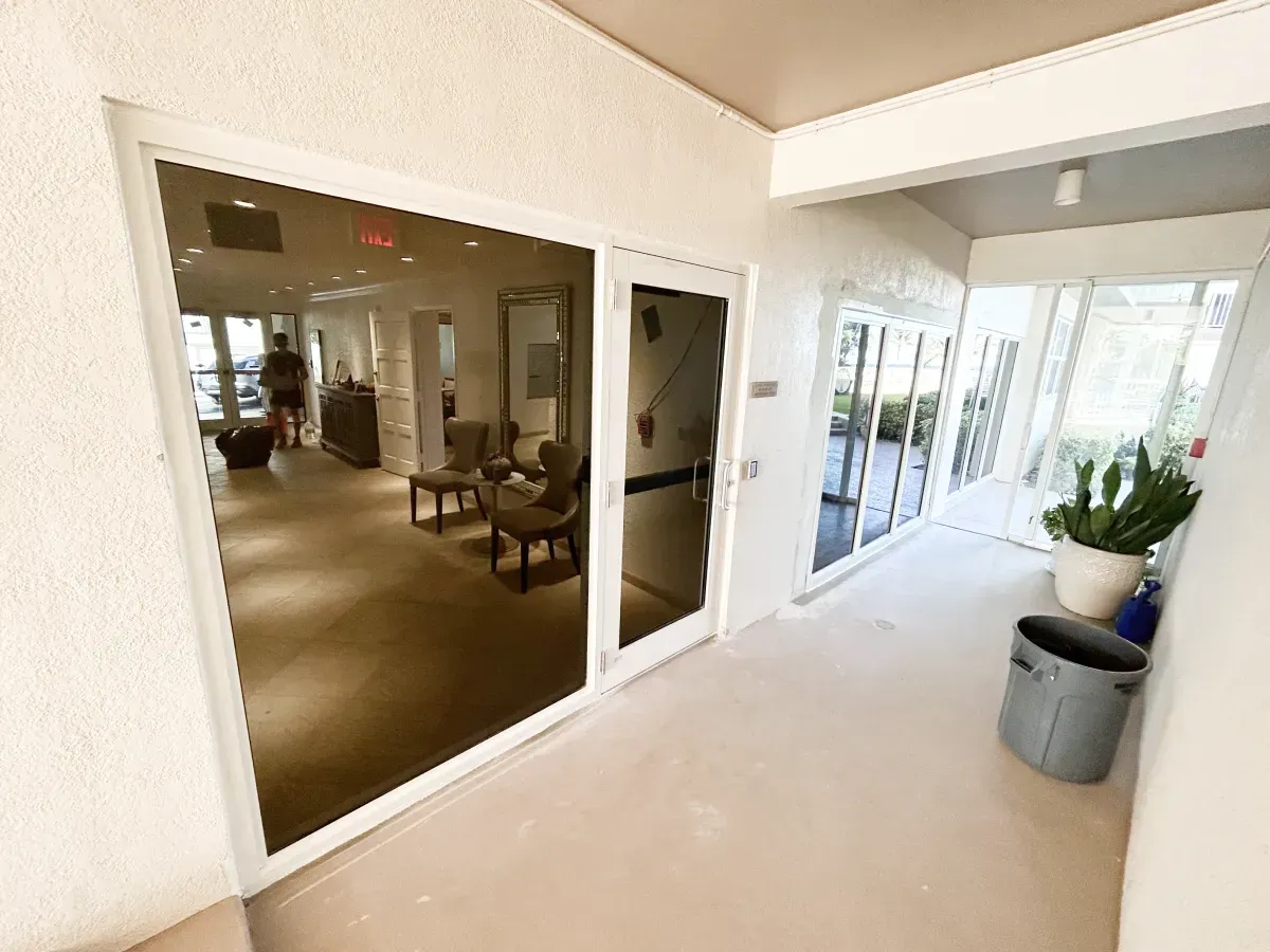 A brightly lit hallway leads to a building entrance with glass doors reflecting an interior lounge area and a potted plant.