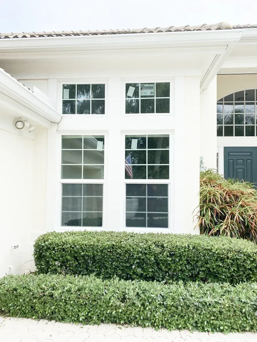 A white exterior wall featuring a double-window setup with gridded panes above a neatly trimmed green hedge.