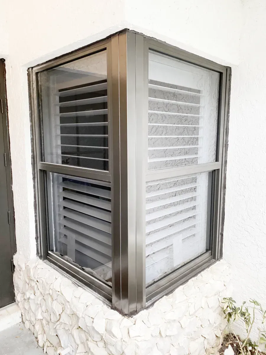 A corner window with a dark metal frame set into a white stone wall, with blinds visible through the glass panes.