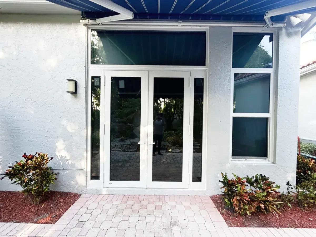 Double glass doors with a rectangular transom and a side window, set in a white stucco wall with landscaping on a patio.