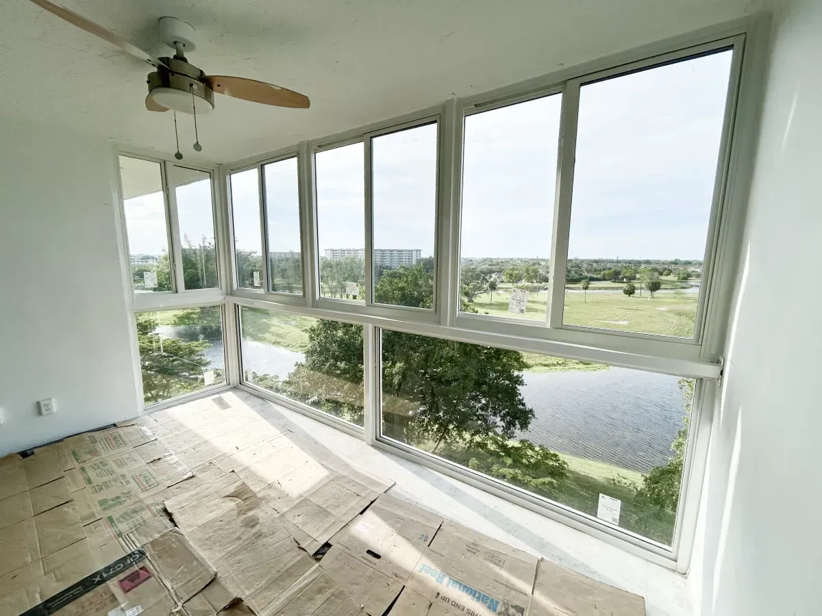 An indoor sunroom with white walls, a ceiling fan, and large windows overlooking a lake and grassy field.