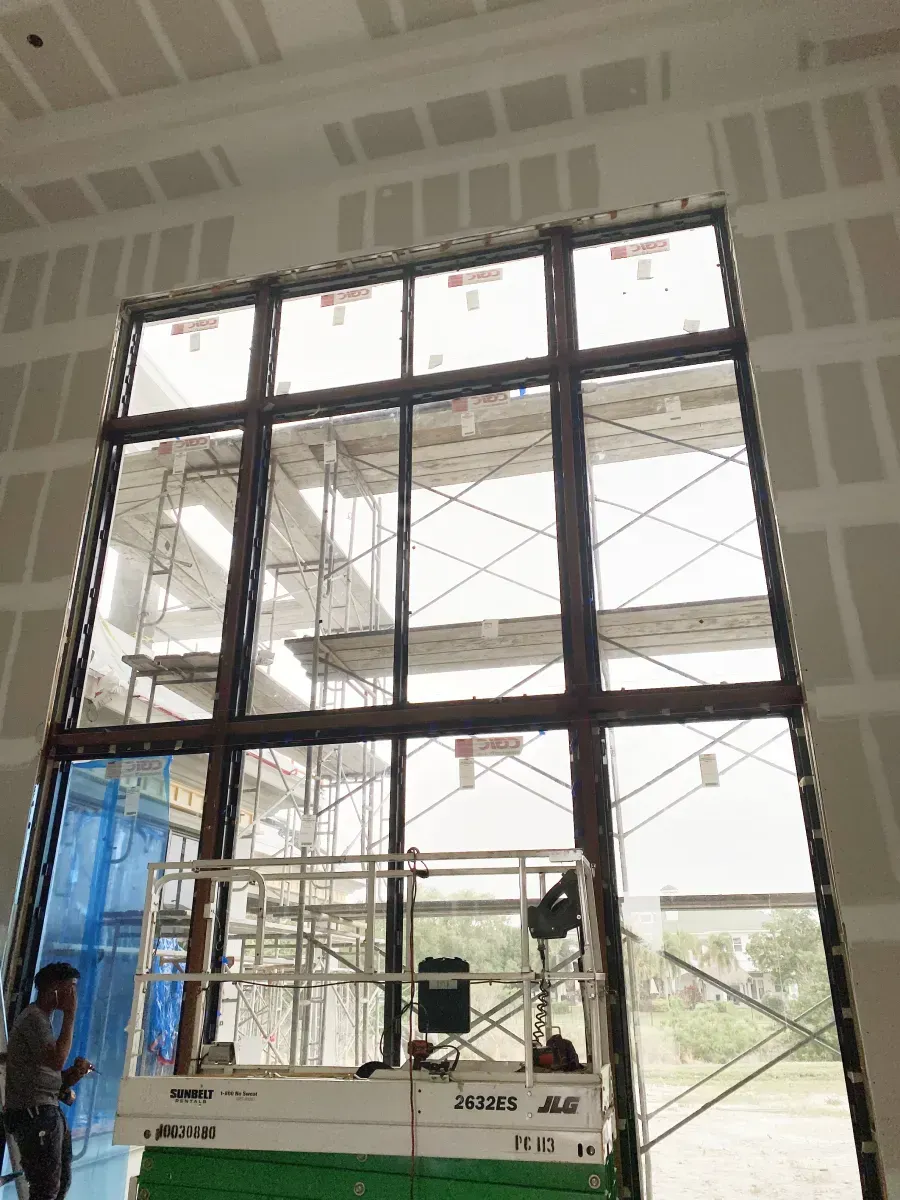 A person standing on an elevated platform works on a large, multi-paned window frame inside a building under construction.