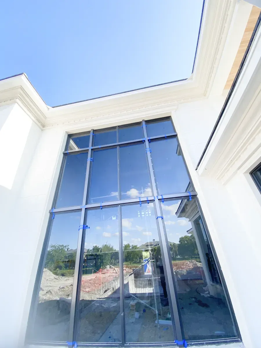 A large, tall glass window on a white exterior wall against a clear blue sky, showing a construction site in the reflection.