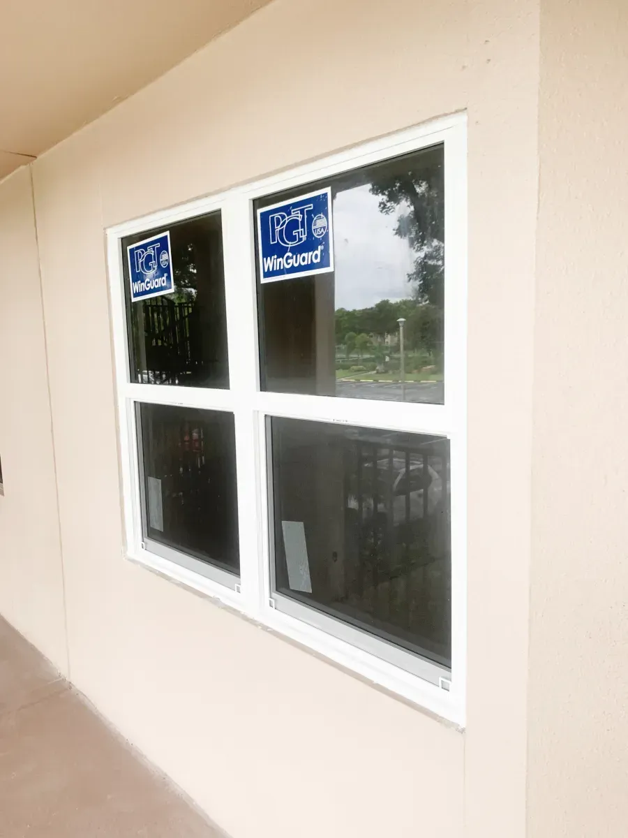 A white-framed window with four panes installed on a beige exterior wall, showing blue manufacturer stickers on the glass.