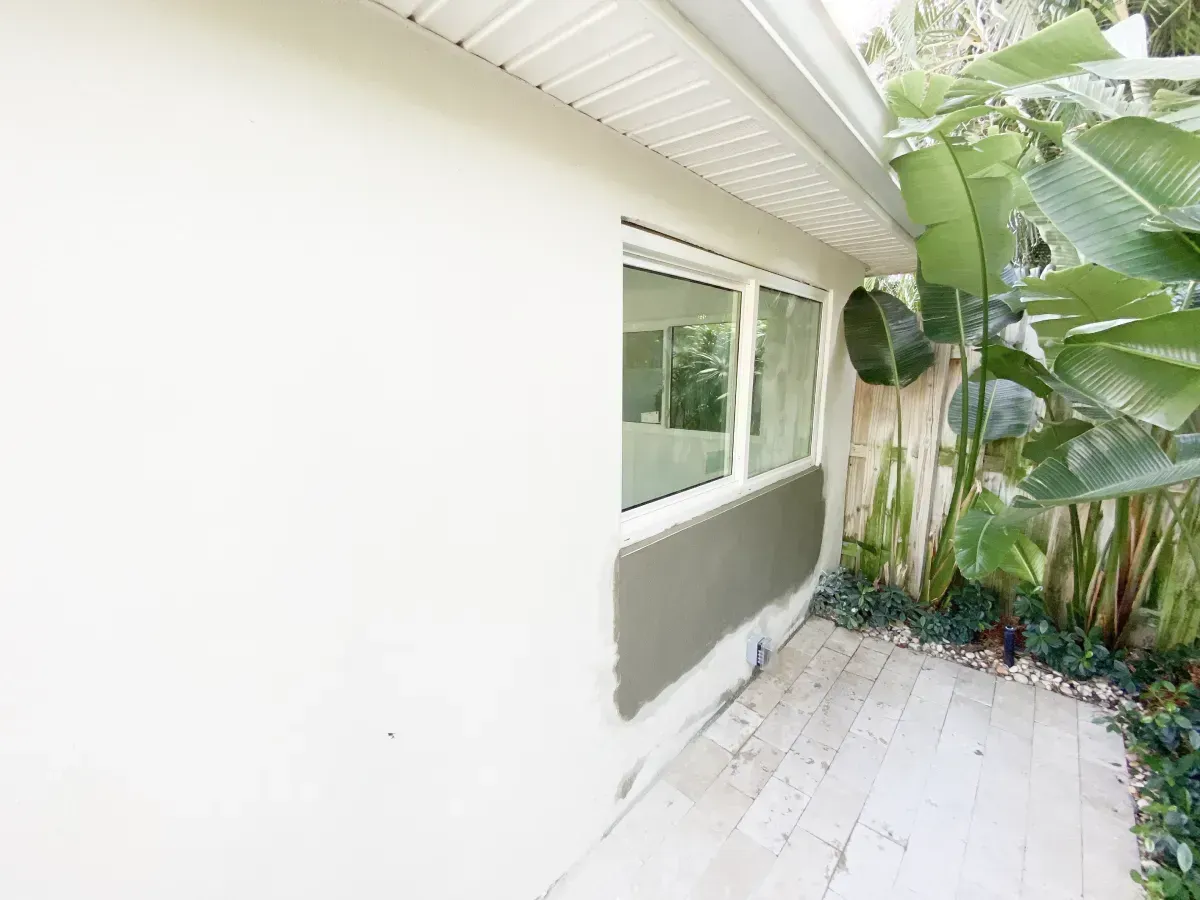 Exterior wall of a light-colored home with a window and a partially applied patch of gray stucco below it.