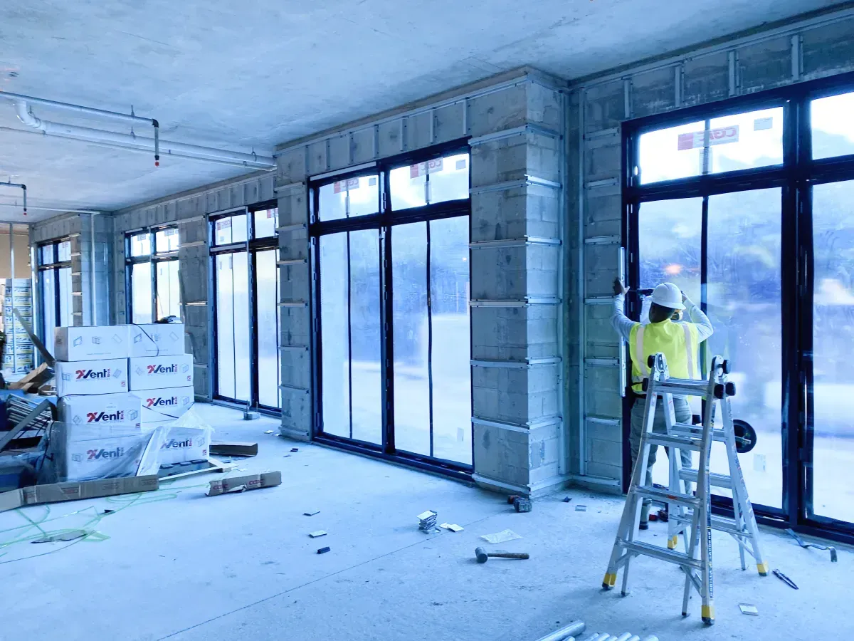 A worker in a yellow safety vest stands on a ladder, installing a large window frame in an unfinished building interior.