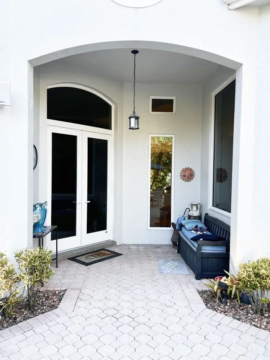 A white-walled home entrance with a double door, a vertical window, a hanging lantern, and a bench on a paved patio.