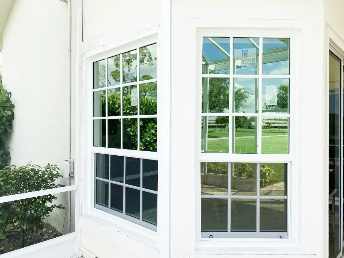 Two white-framed, double-hung windows with a grid pattern, reflecting a green lawn and trees on their glass panes.