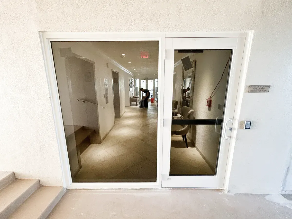 White double glass doors lead into a bright, tiled lobby with interior lighting, viewed from a small set of concrete steps.