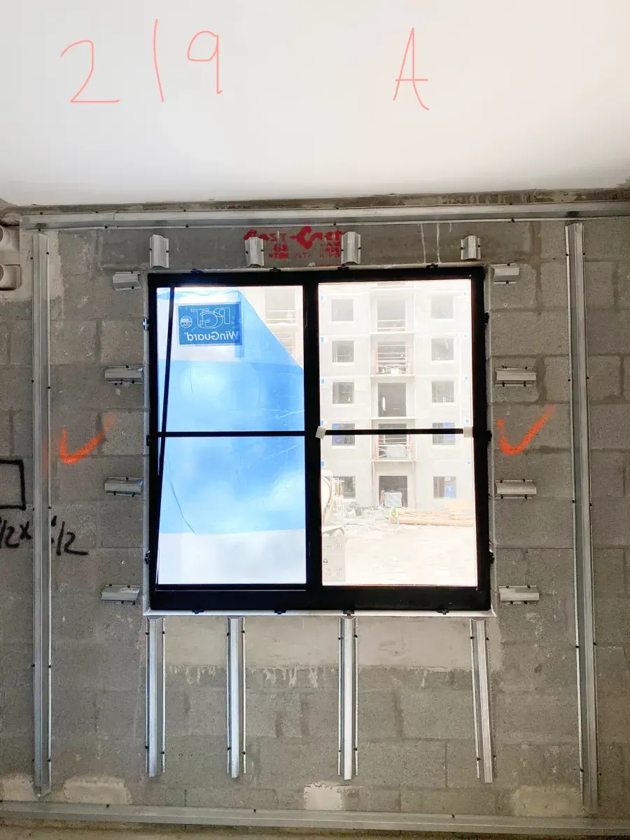 A window frame is installed in a grey concrete block wall, secured with metal brackets and vertical supports.