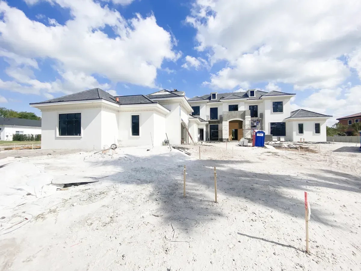 A large, white, modern house under construction with a dark roof and unfinished sandy yard under a blue sky.