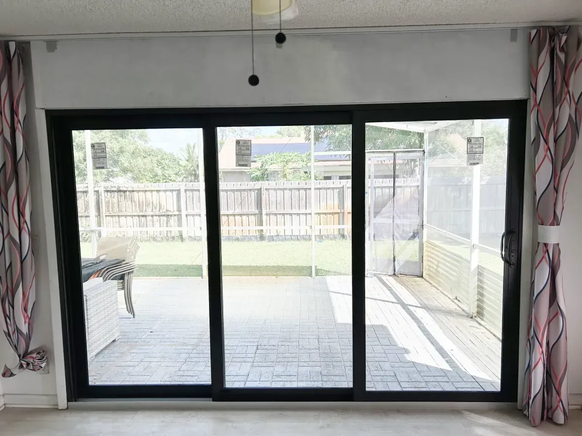 Large sliding glass patio doors with a black frame, flanked by patterned curtains, looking out onto a paved backyard patio.