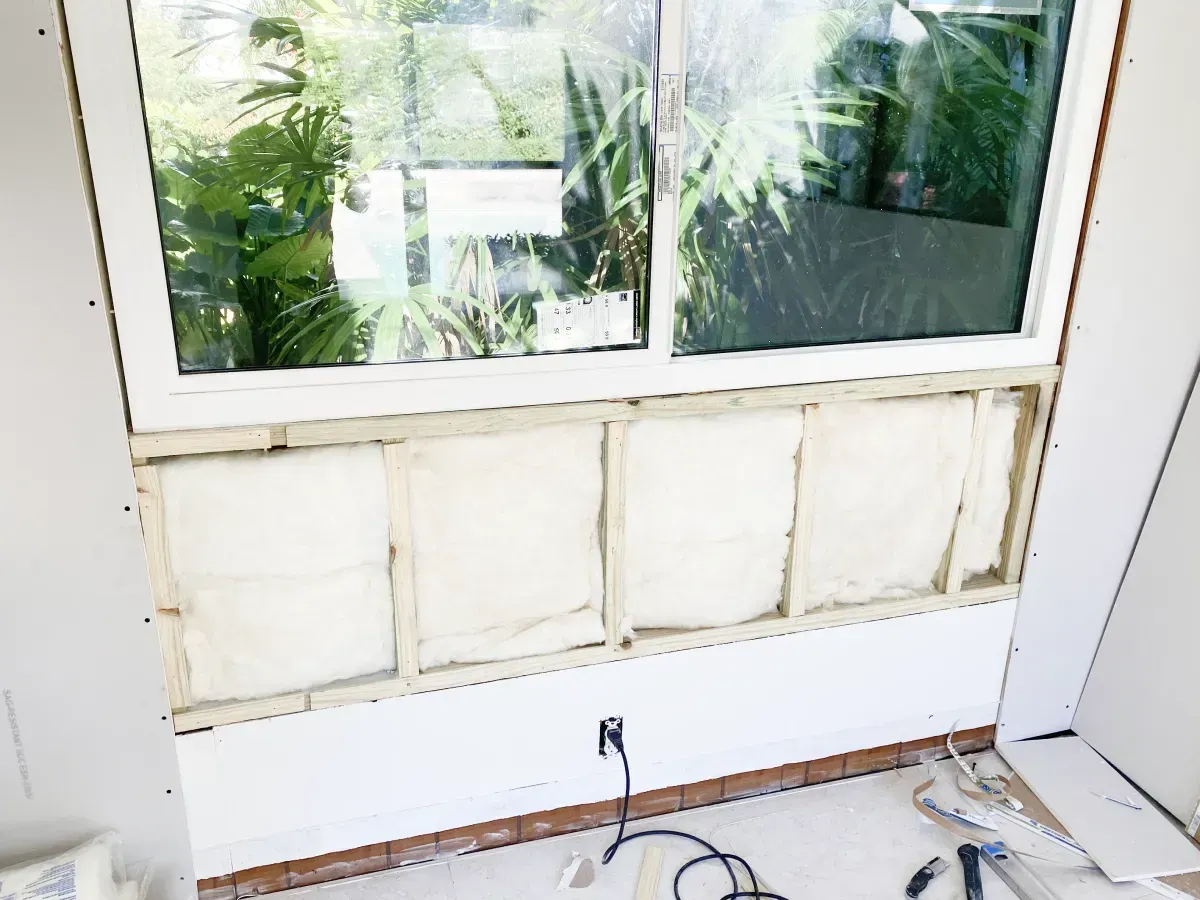 A wall section under a window is framed with wood studs and filled with white insulation, with drywall nearby and tools.