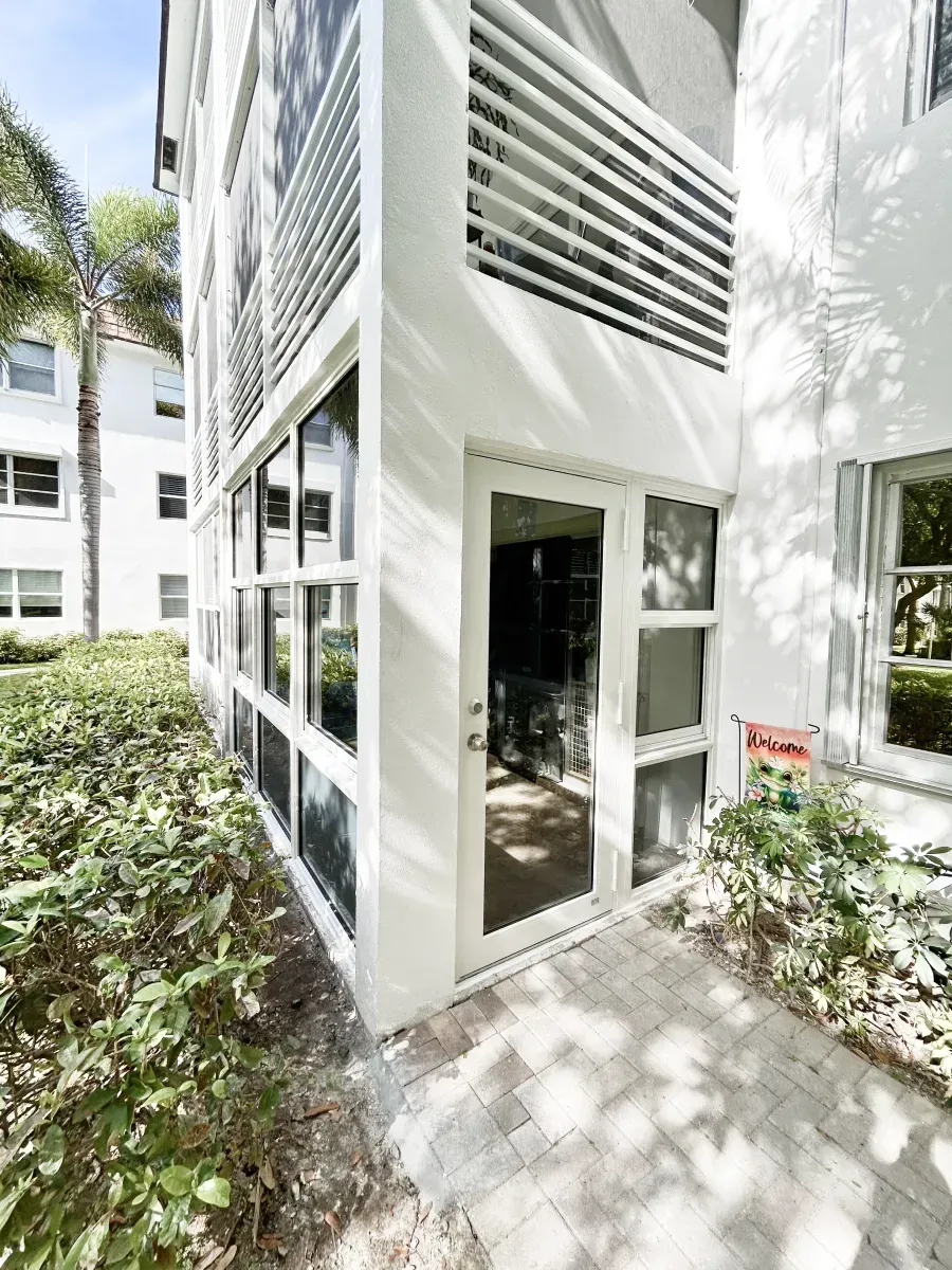 A white residential building exterior with a glass door, screened-in porch, and a brick walkway surrounded by greenery.