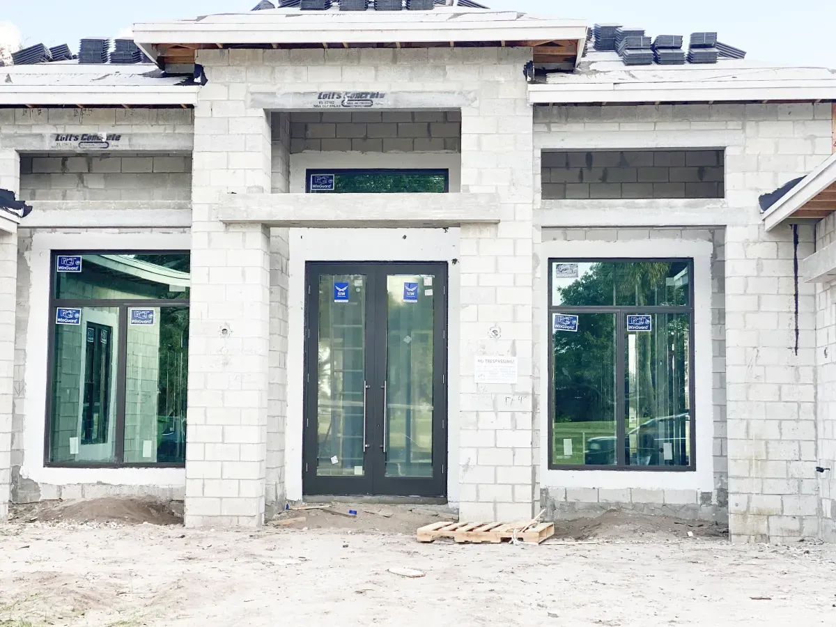 Construction site showing the front facade of a concrete block house with a central double door and two large windows.
