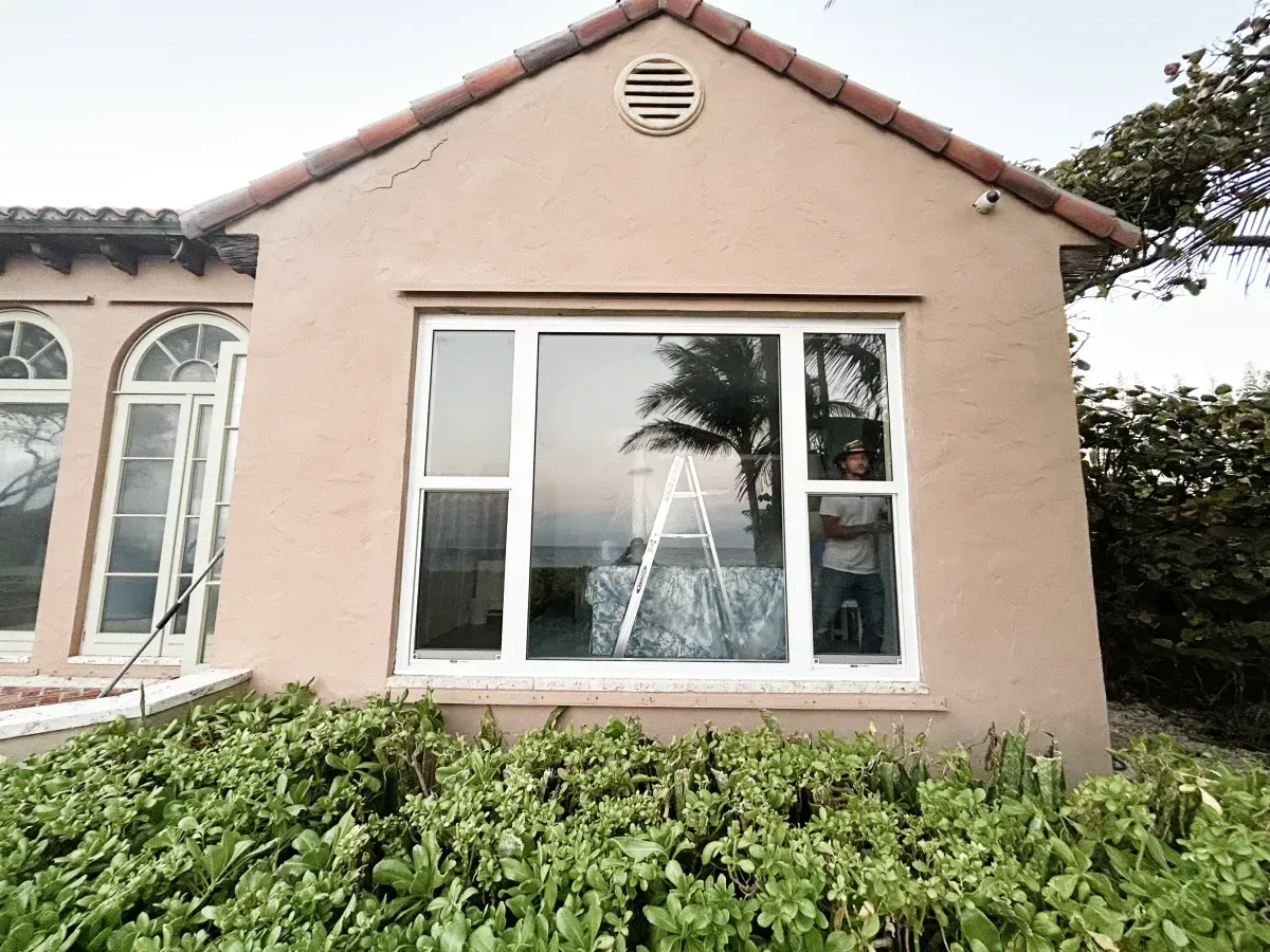 A person stands inside a room viewed through a large window on the front of a tan house with a ladder behind them.