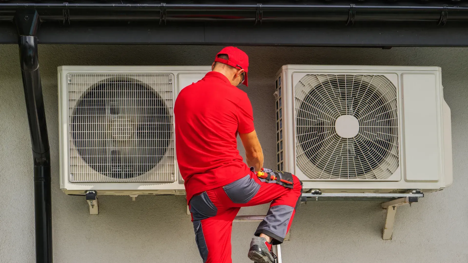 A man is standing on a ladder fixing two air conditioners on the side of a building.