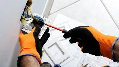 A person wearing orange and black gloves is using a screwdriver to fix an electrical outlet.